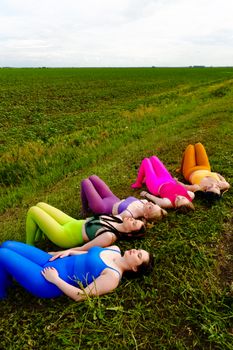 Group of women in vibrant outfits lying on grass in an open field under a clear sky.