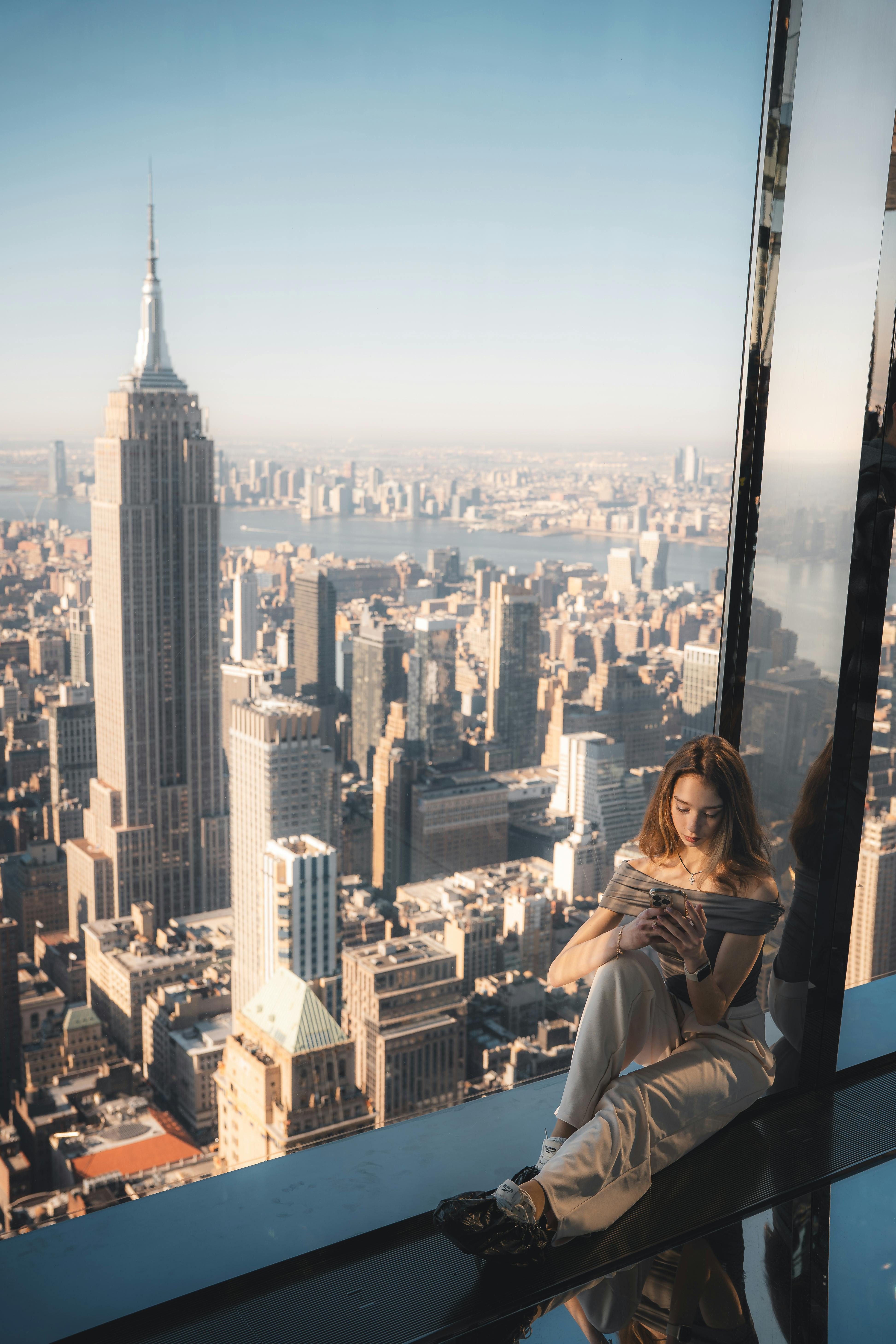 A woman sitting by a window with a stunning view of New York City's skyline and the Empire State Building.