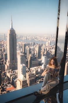 A woman sitting by a window with a stunning view of New York City's skyline and the Empire State Building.