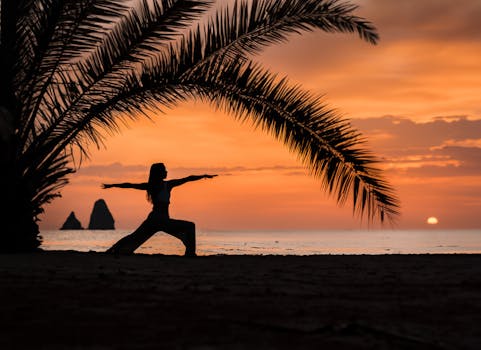 A woman practices yoga under palm trees during a vibrant sunset on a tropical beach.
