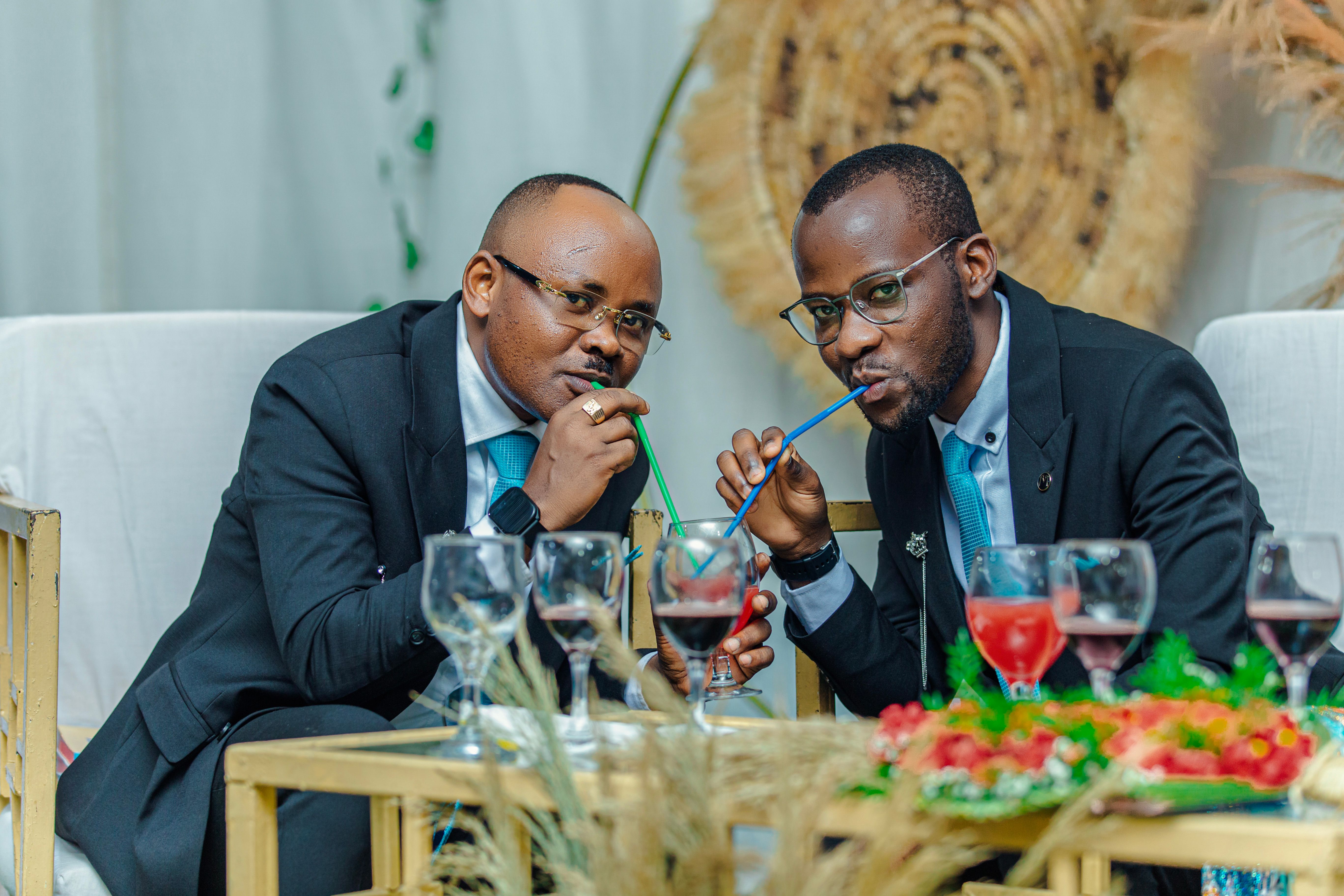 Two elegantly dressed men in suits enjoying a drink with straws at a social event.