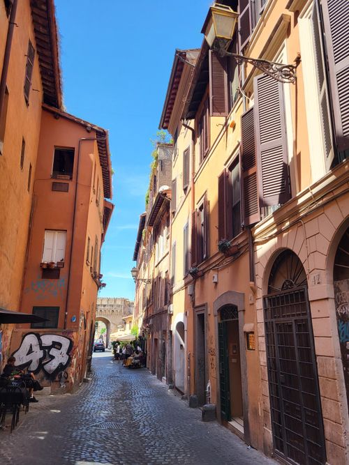Free Explore the narrow, sunlit streets of Trastevere, Rome, featuring vibrant facades and cobblestone paths. Stock Photo