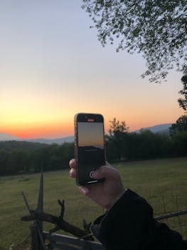 A hand holding a smartphone capturing a beautiful outdoor sunset scene.