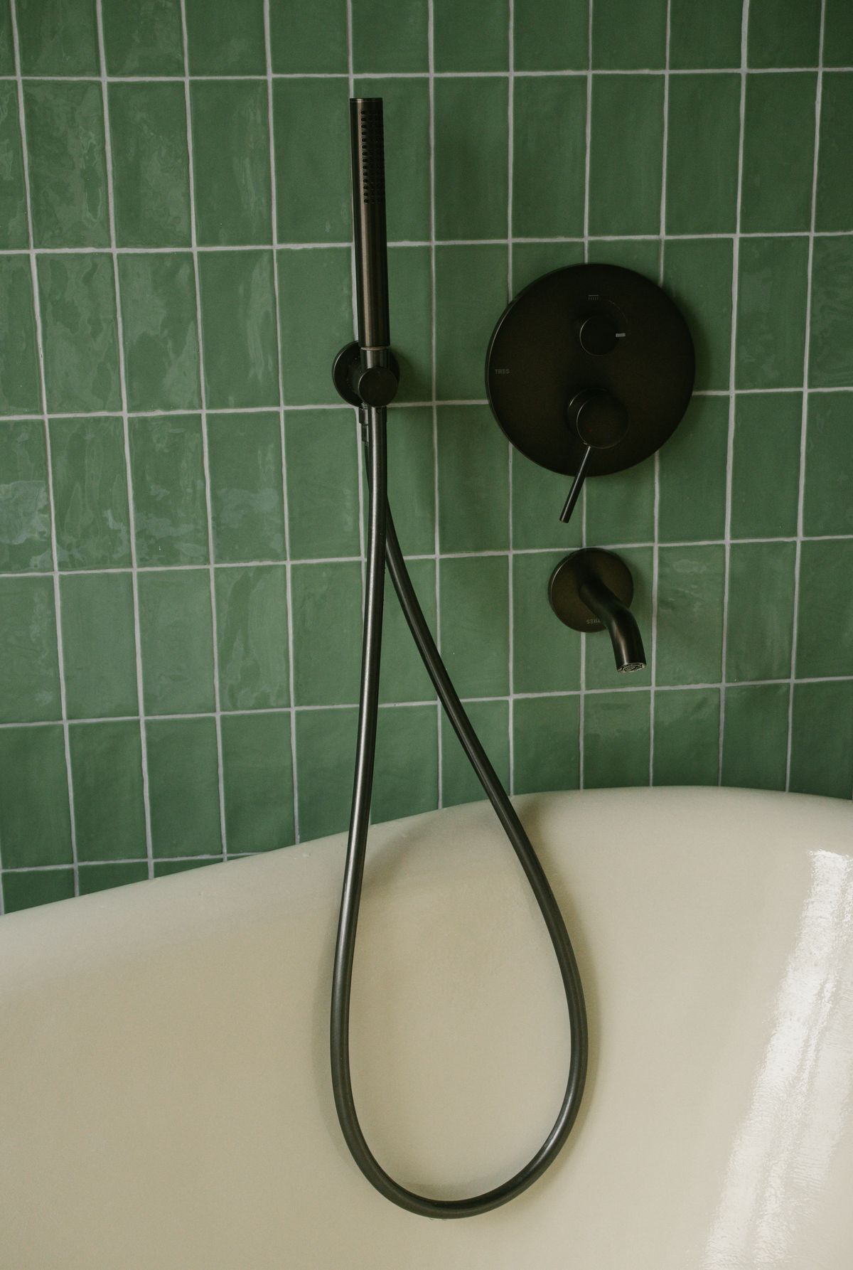 Close-up of a bathtub with green square wall tiles, dark grout and matte-black shower fittings.