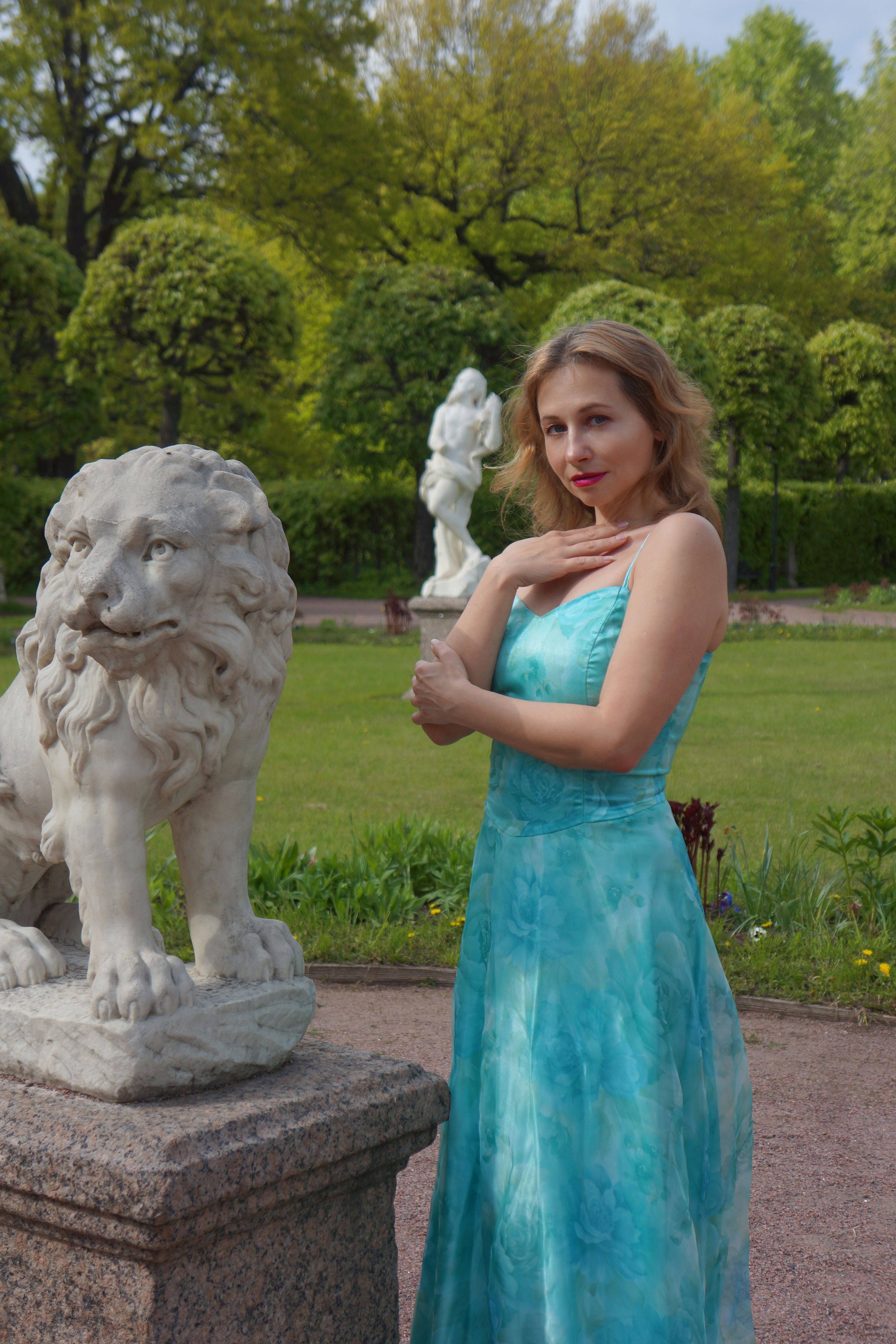 A woman in a turquoise dress poses beside marble statues in a serene garden setting.