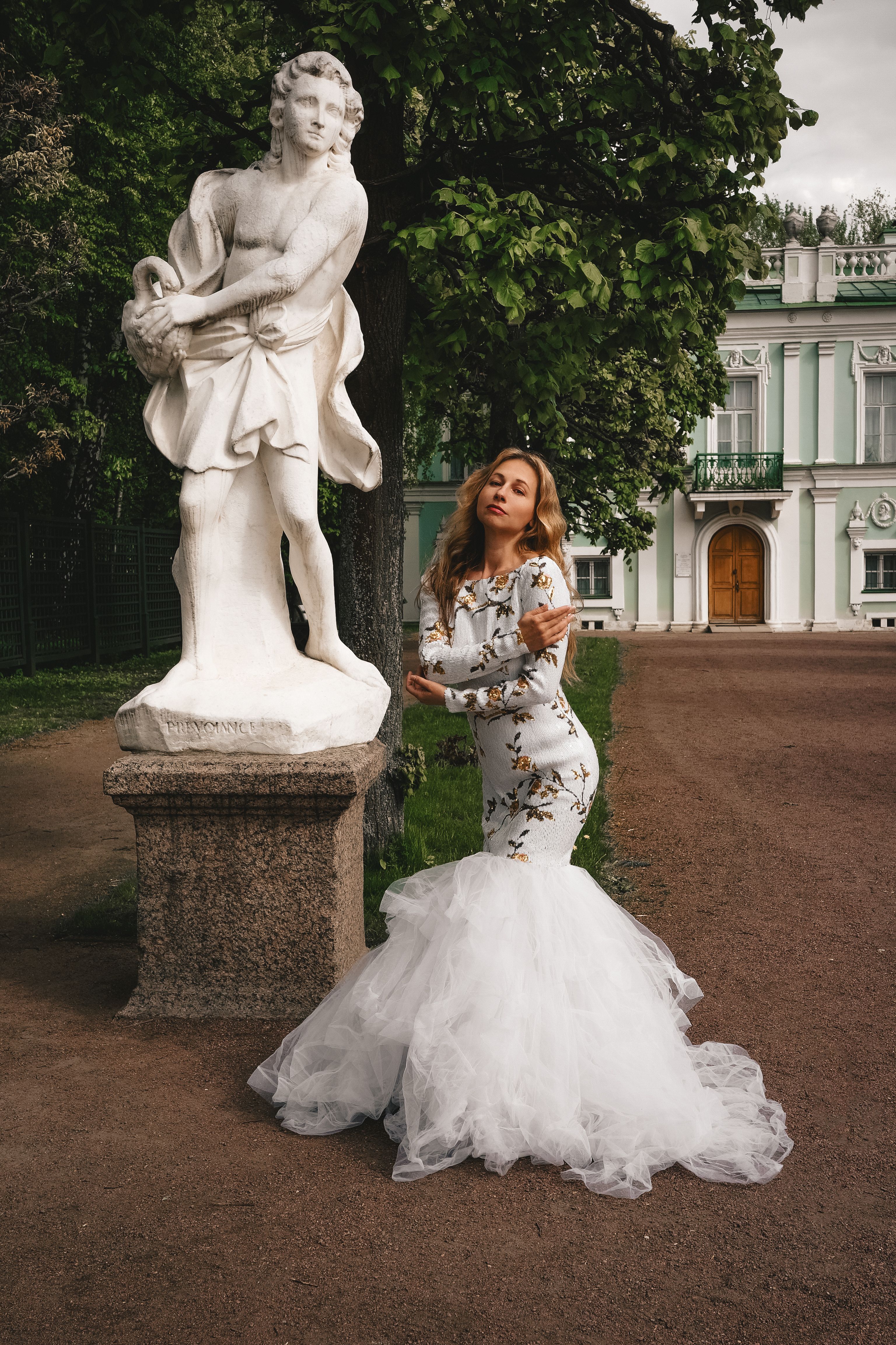 A woman in a white dress poses gracefully beside a classical statue in a garden setting.
