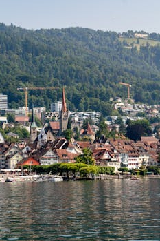 Picturesque view of Zug town, Switzerland, by the lake with lush hills and vibrant rooftops under a clear summer sky.