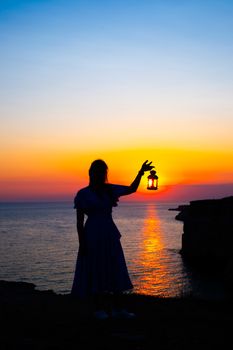 A stunning silhouette of a person holding a lantern by the sea during sunset, creating a dramatic scene.