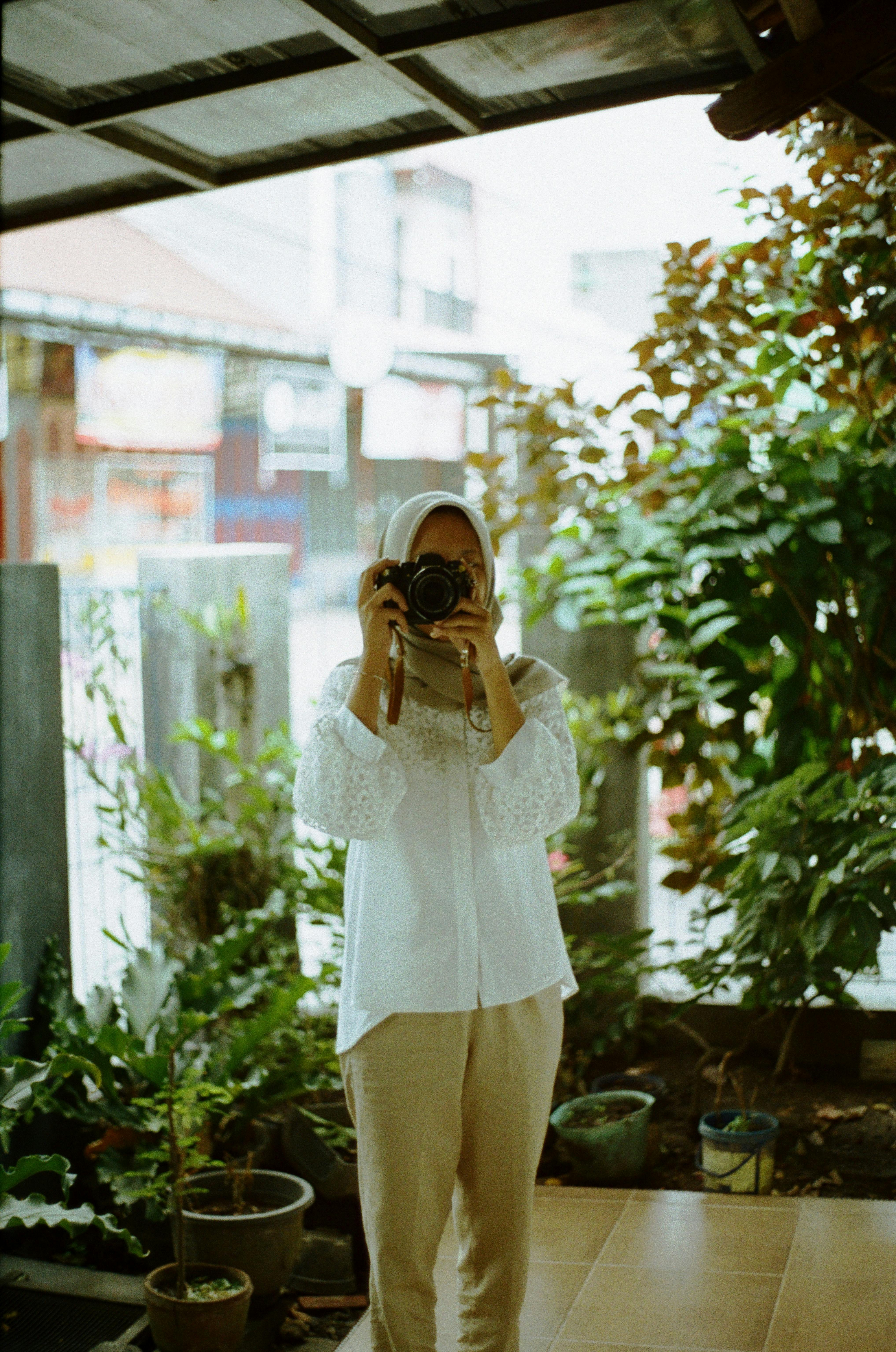 A woman stands indoors with a camera, surrounded by lush plants, capturing the vibrant scene.