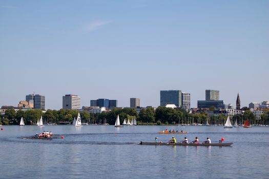 Rowers and sailboats on Alster River in Hamburg on a sunny summer day with city skyline.