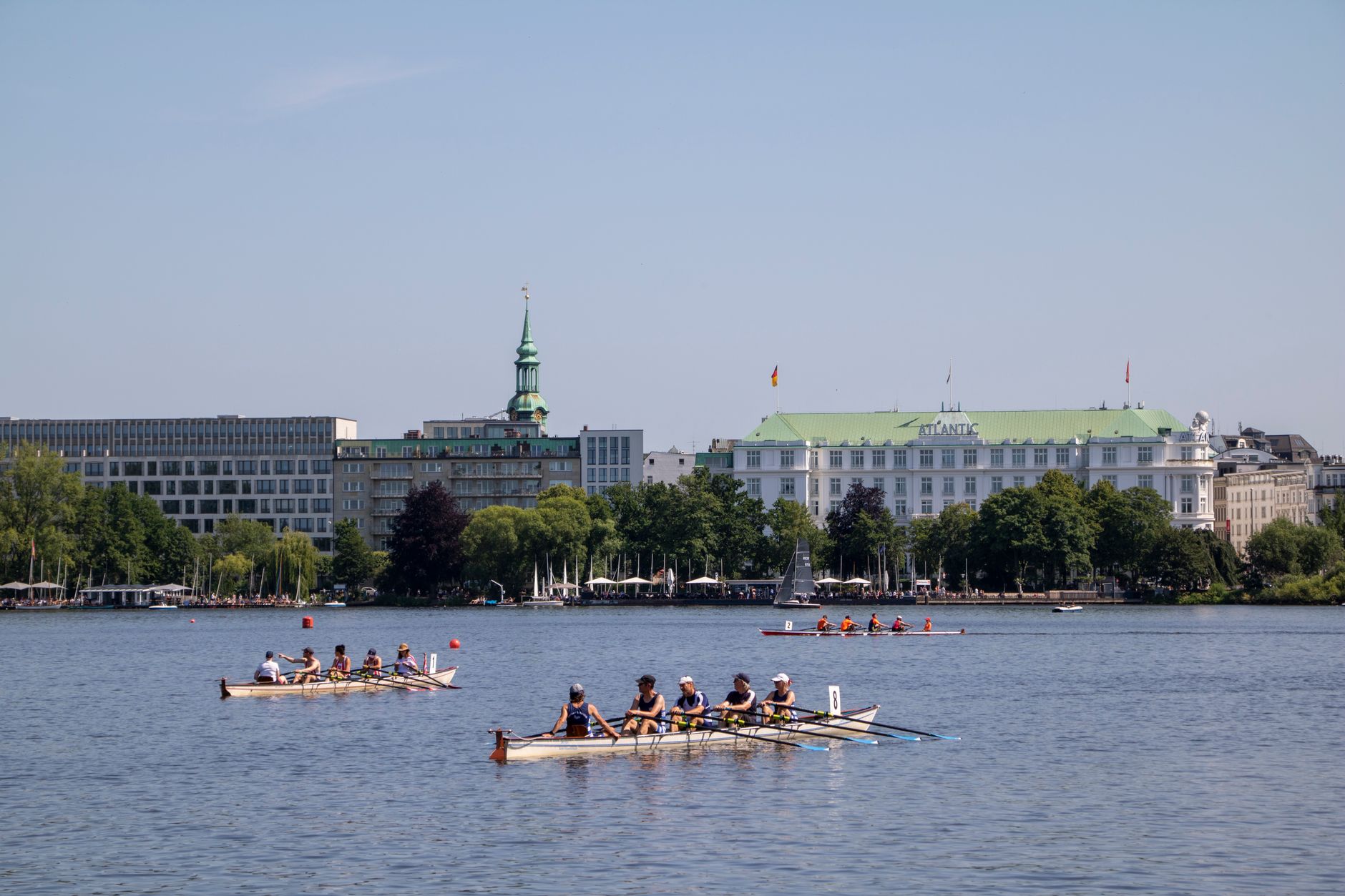 Rowing teams glide on Alster River with Hamburg's architectural skyline in the background under a clear summer sky.