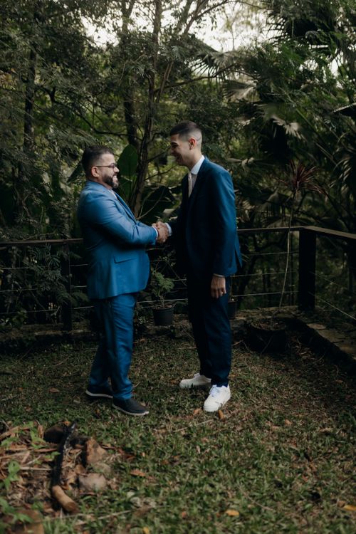 Free Two men in formal suits engaged in a handshake amidst lush greenery. Stock Photo