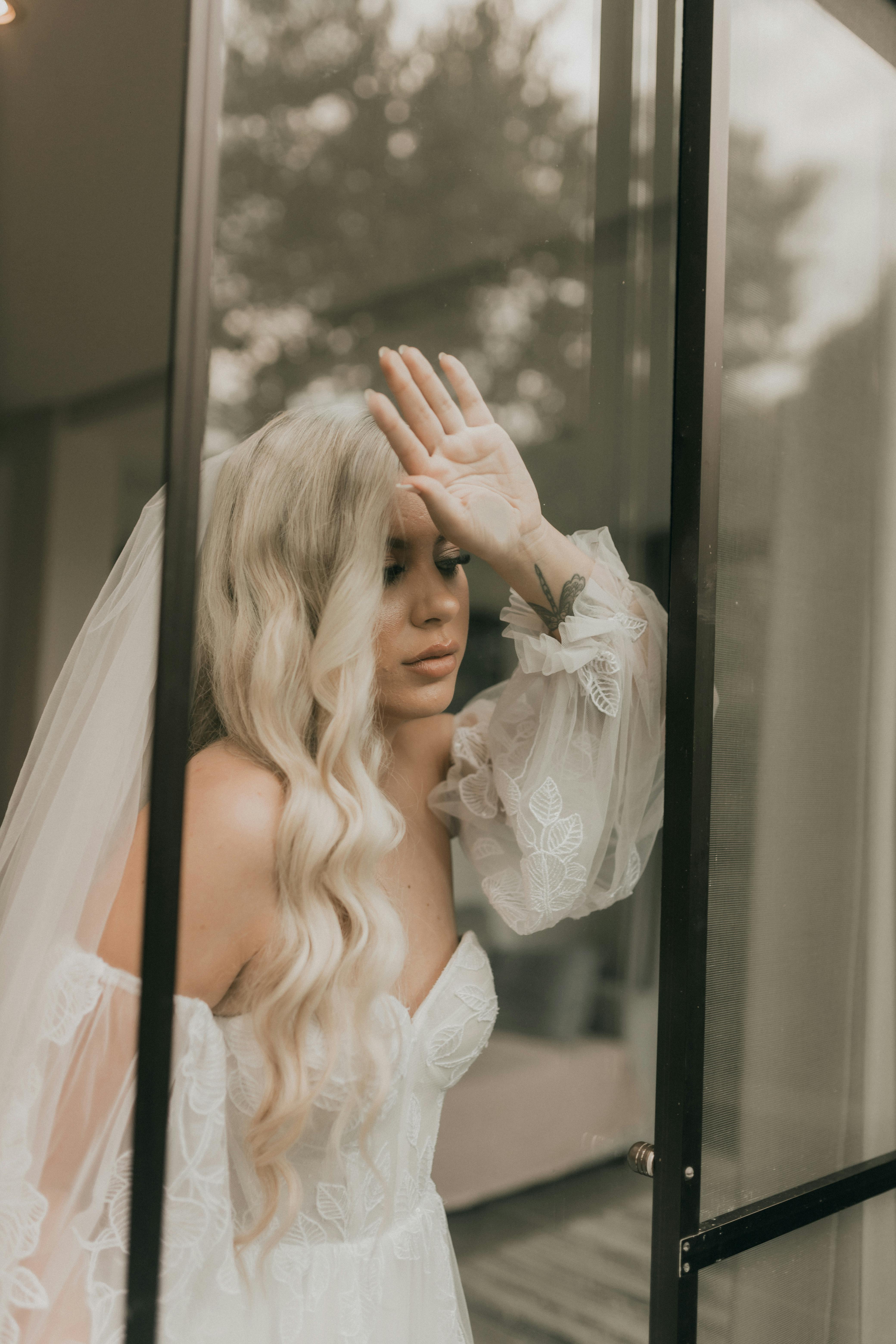 Bride in white gown and veil gazes thoughtfully through a window, bathed in natural light.
