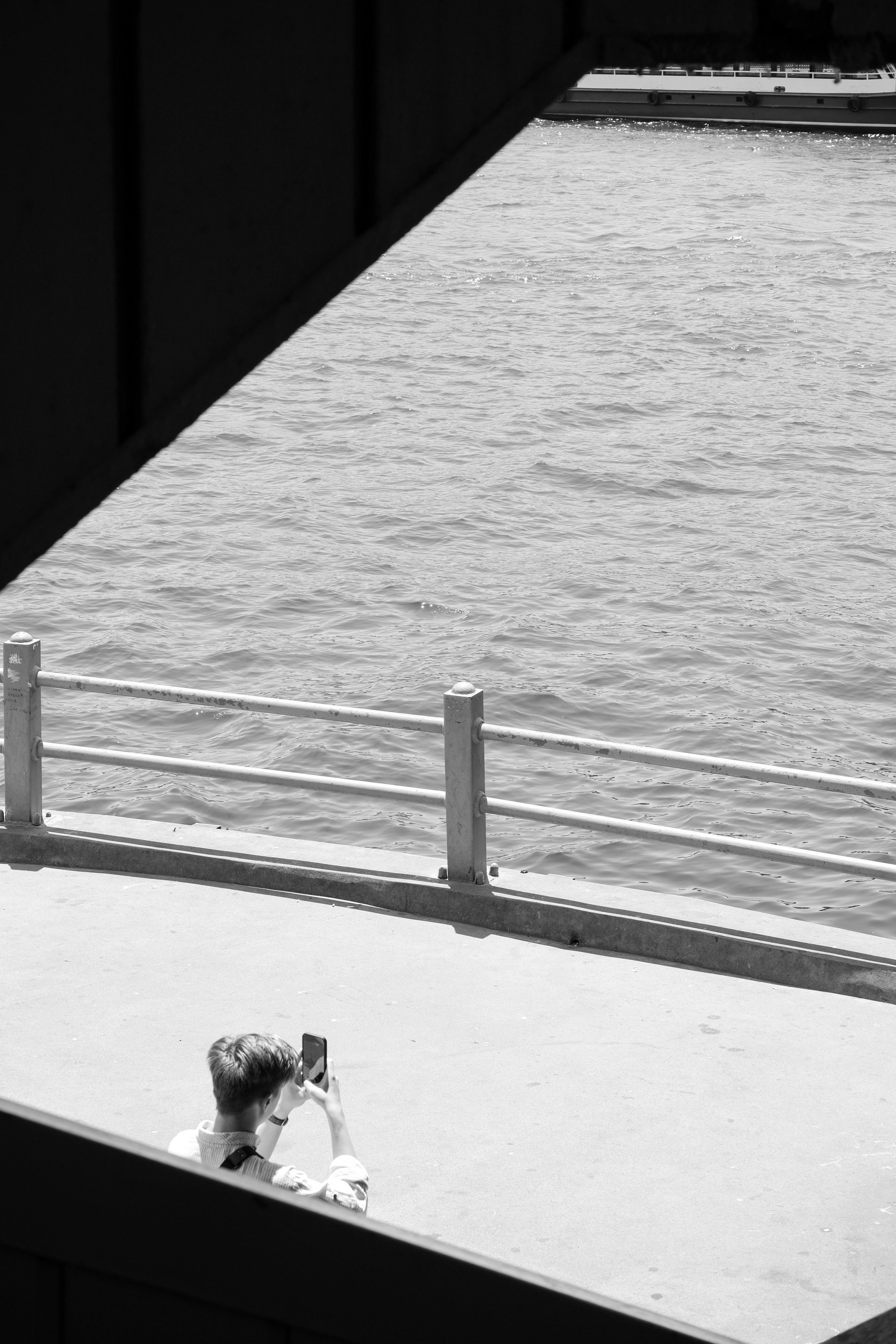 Free A solitary figure captures a waterfront view on a smartphone, framed in black and white. Stock Photo