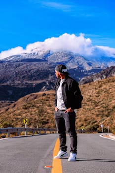 A man stands on a mountain road gazing at snowy peaks under a clear sky.