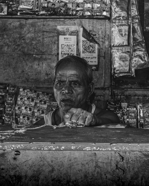 Free Black and white photo of an elderly vendor in a traditional market stall with packaged goods. Stock Photo