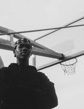 Black and white portrait of a young man standing by a basketball hoop outdoors.