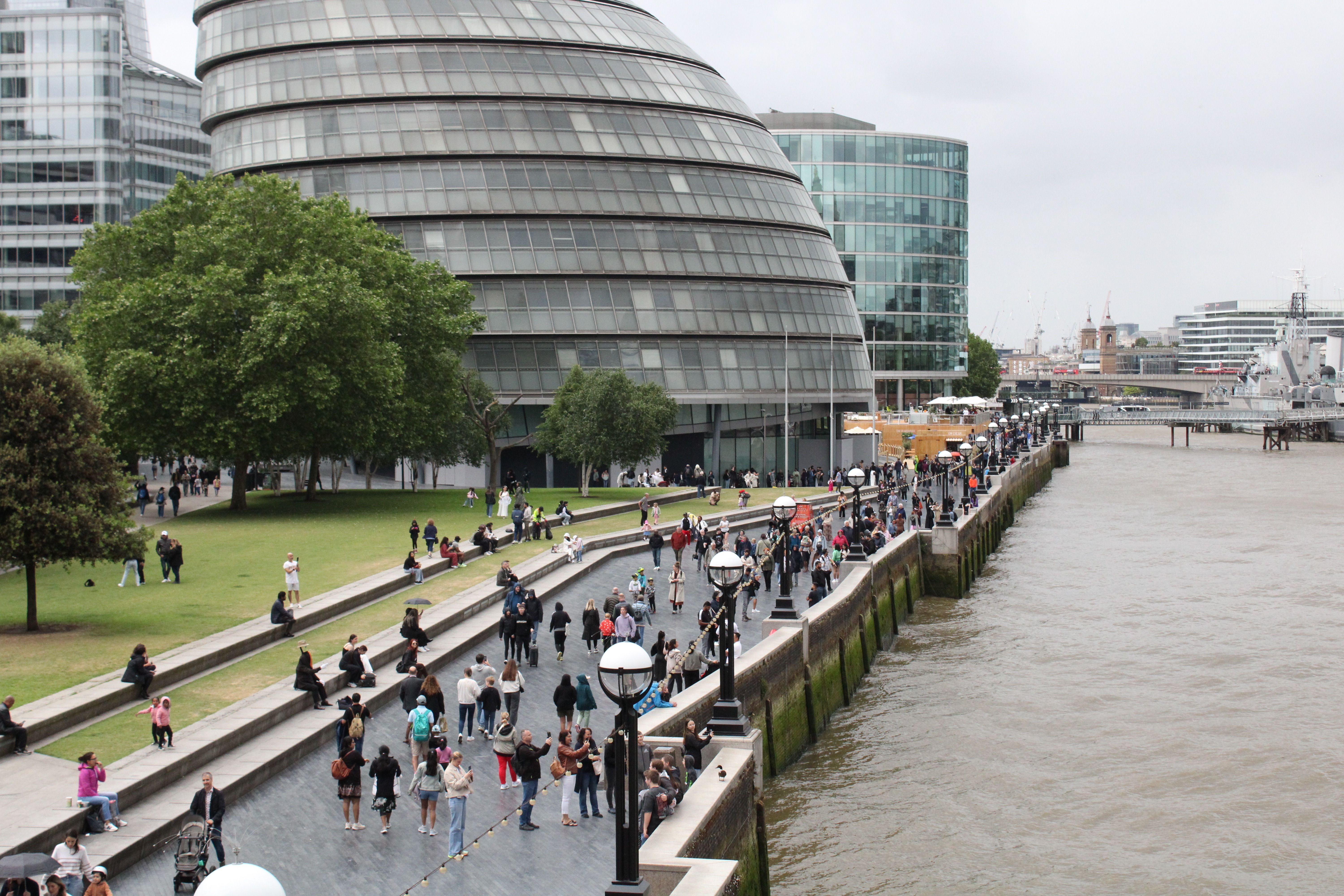 People walking along the waterfront near London's iconic City Hall building on a cloudy day.