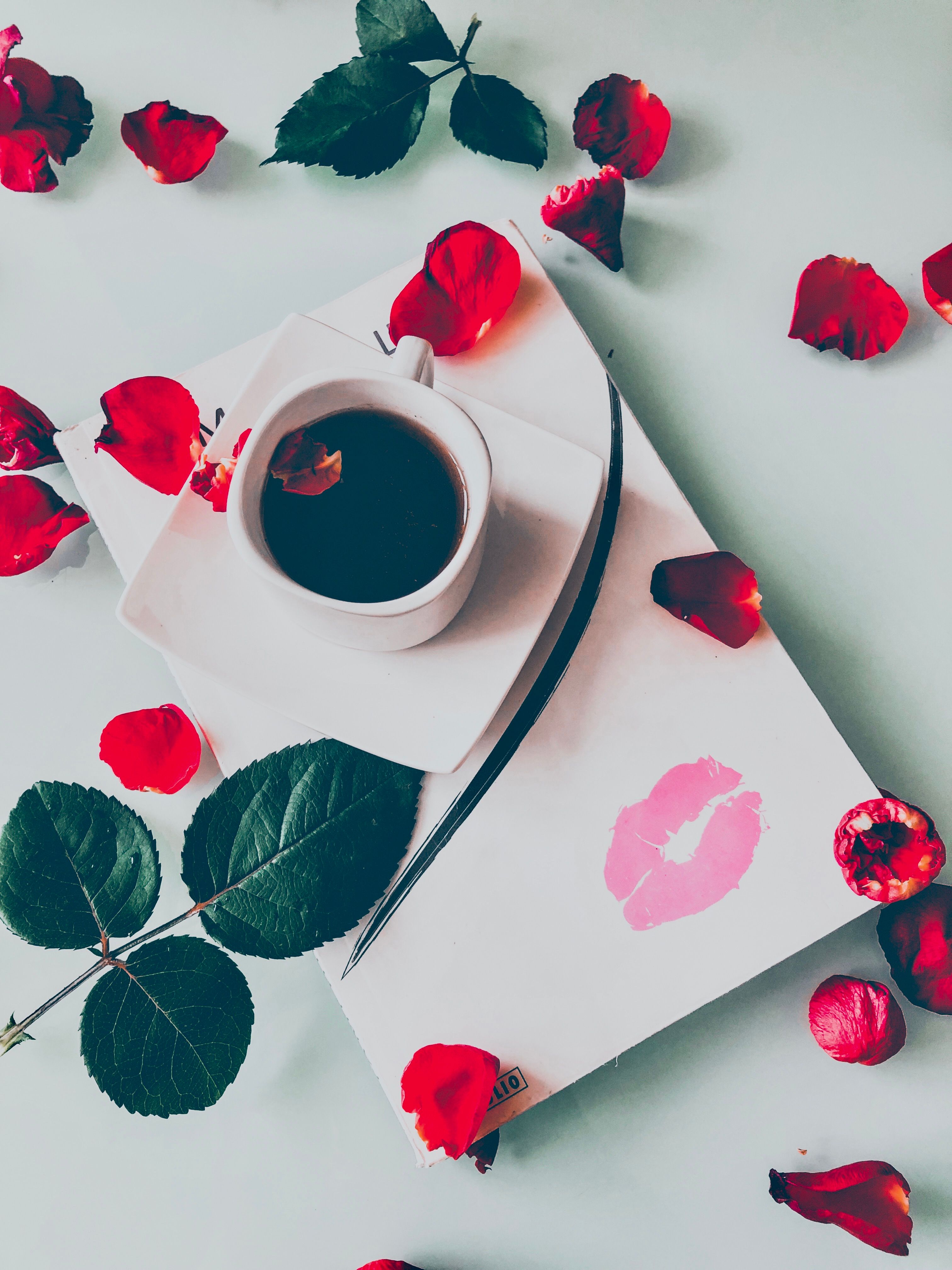 Coffee cup surrounded by red rose petals on white paper with lipstick kiss print.