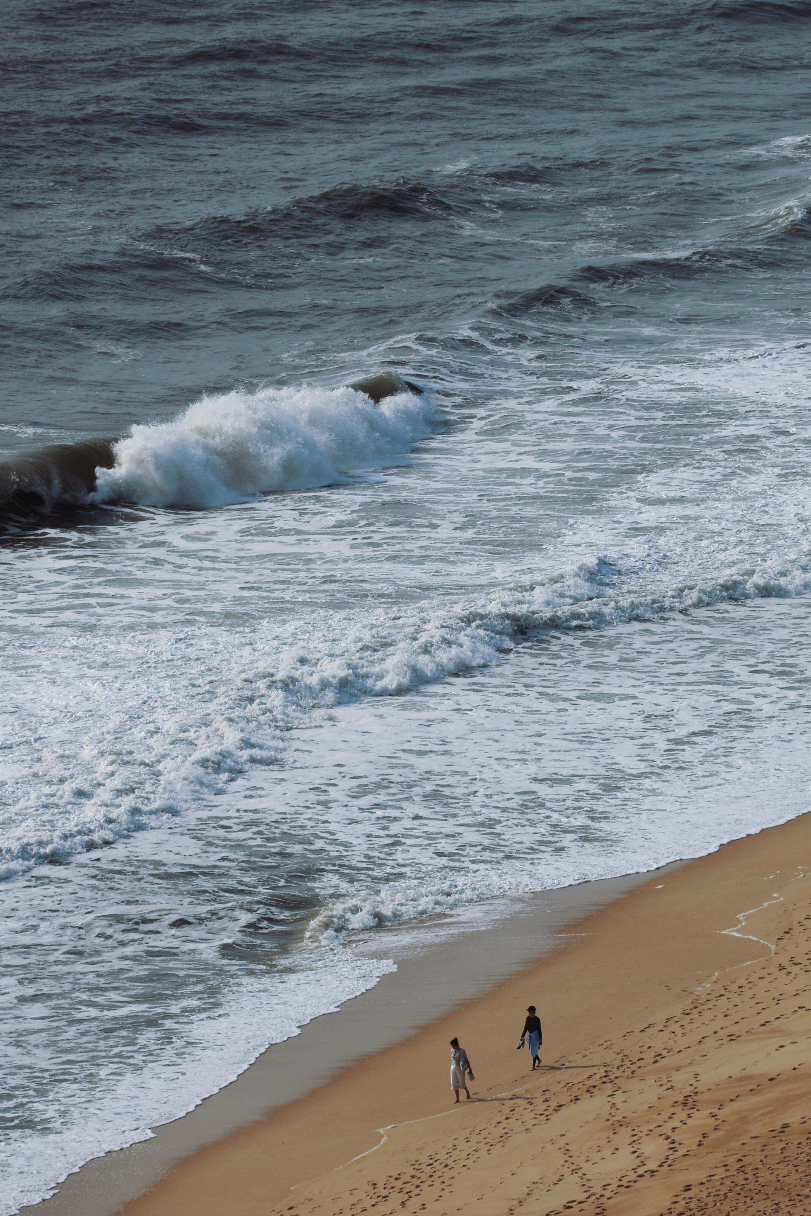 A couple strolls along a sandy beach with waves crashing beside them.