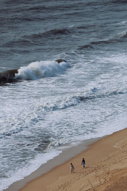 A couple strolls along a sandy beach with waves crashing beside them.