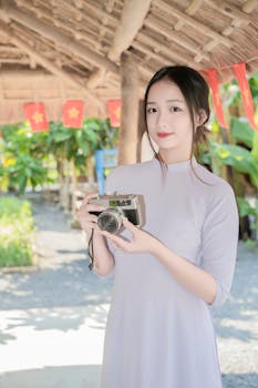 A young woman in traditional clothing holds a vintage camera under a thatched roof.