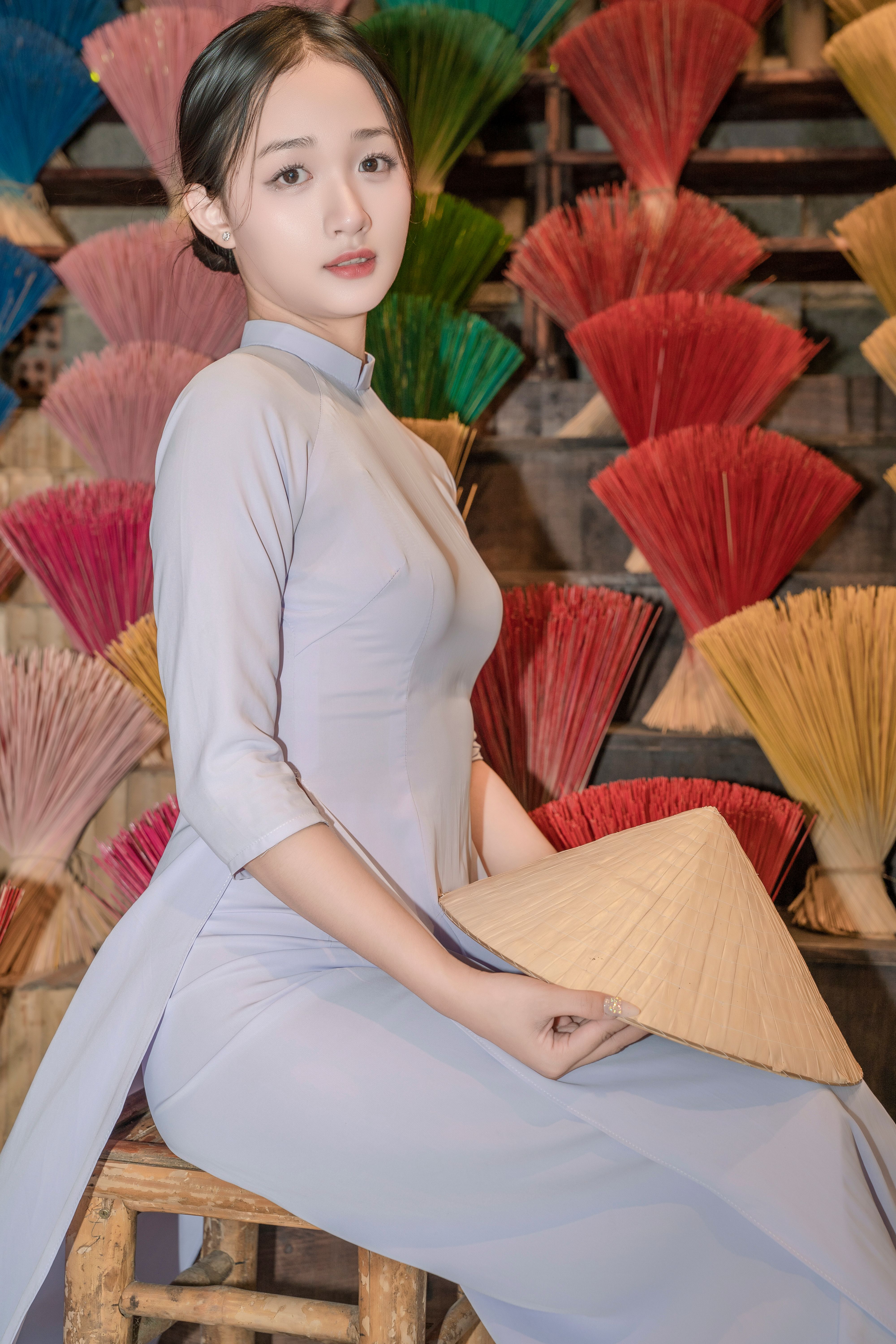 Woman in an elegant áo dài holding a conical hat against a backdrop of colorful incense sticks.