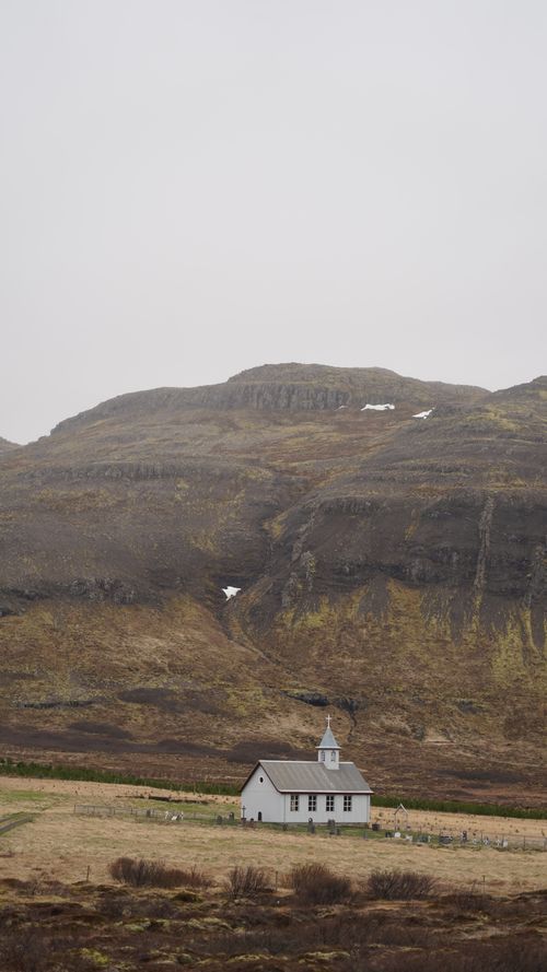 Free A quaint white church nestled in Iceland's rugged, mountainous landscape under a gray sky. Stock Photo