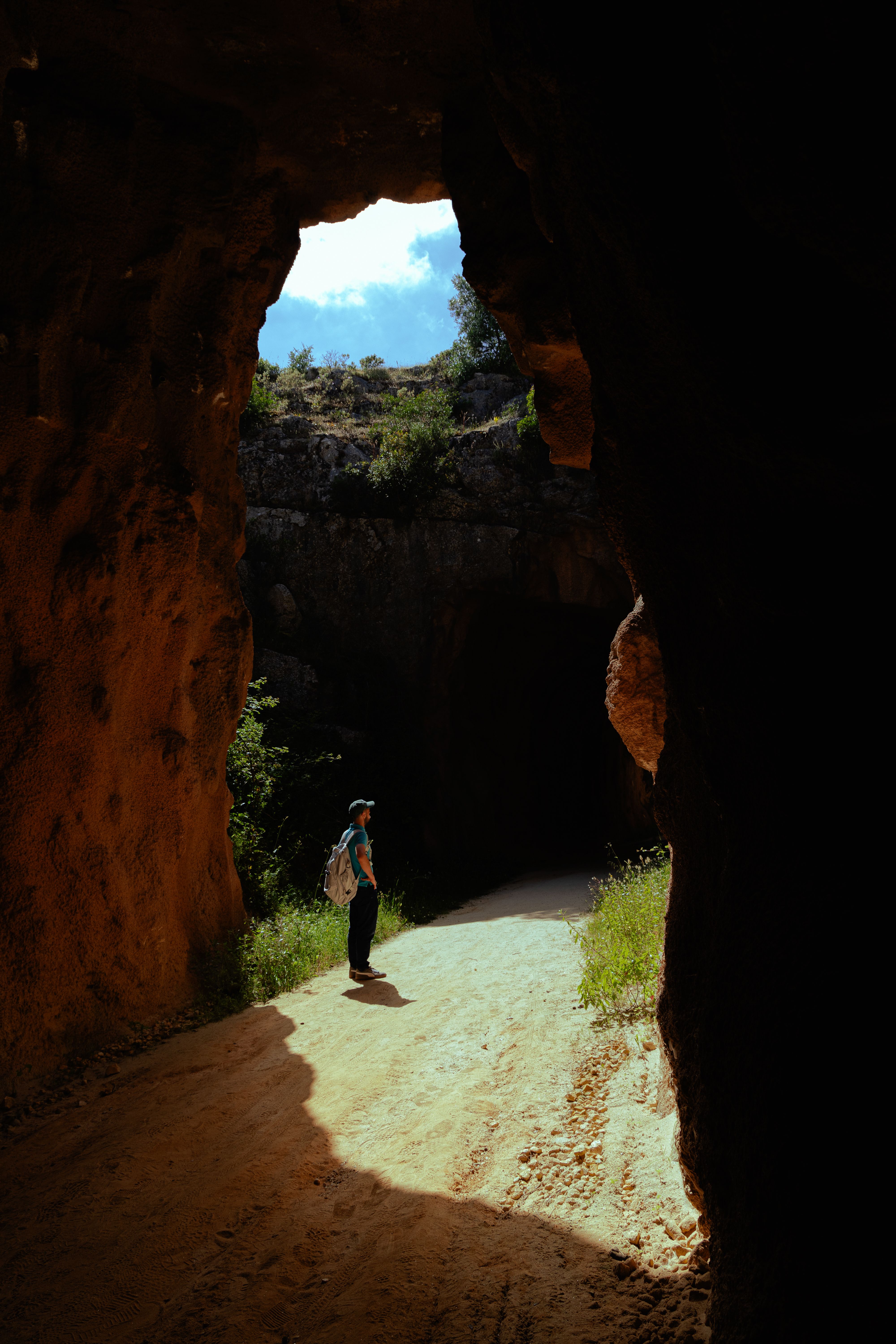 A lone hiker explores a rugged, sunlit cave with vibrant vegetation.