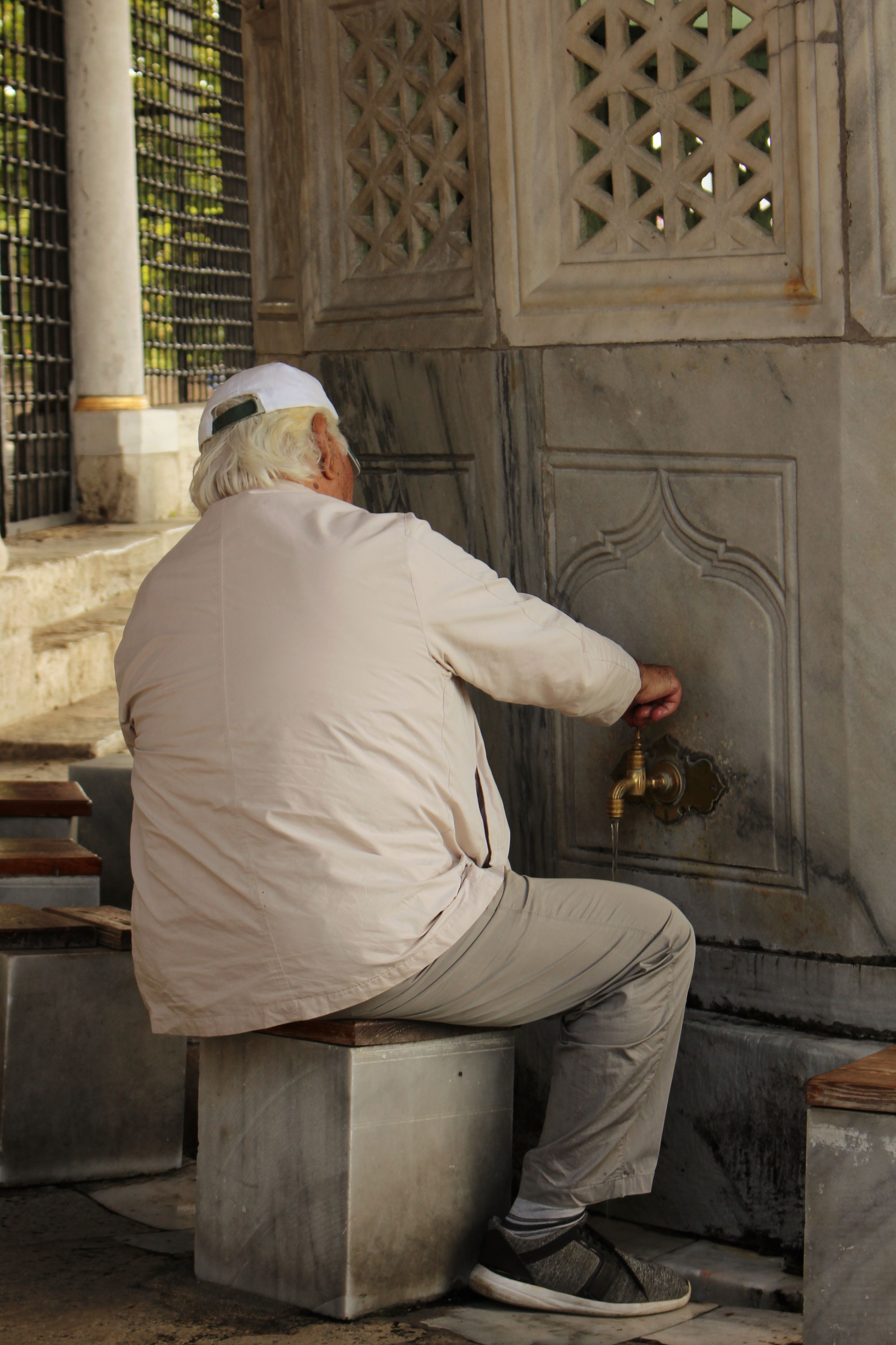 A senior man seated by a traditional water faucet outdoors.