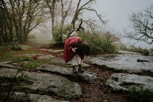 A person in a red coat bends down on a misty forest trail, capturing nature's beauty.