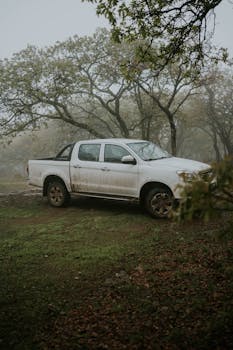 Muddy white pickup truck parked in a foggy forest, evoking a rugged outdoor adventure theme.