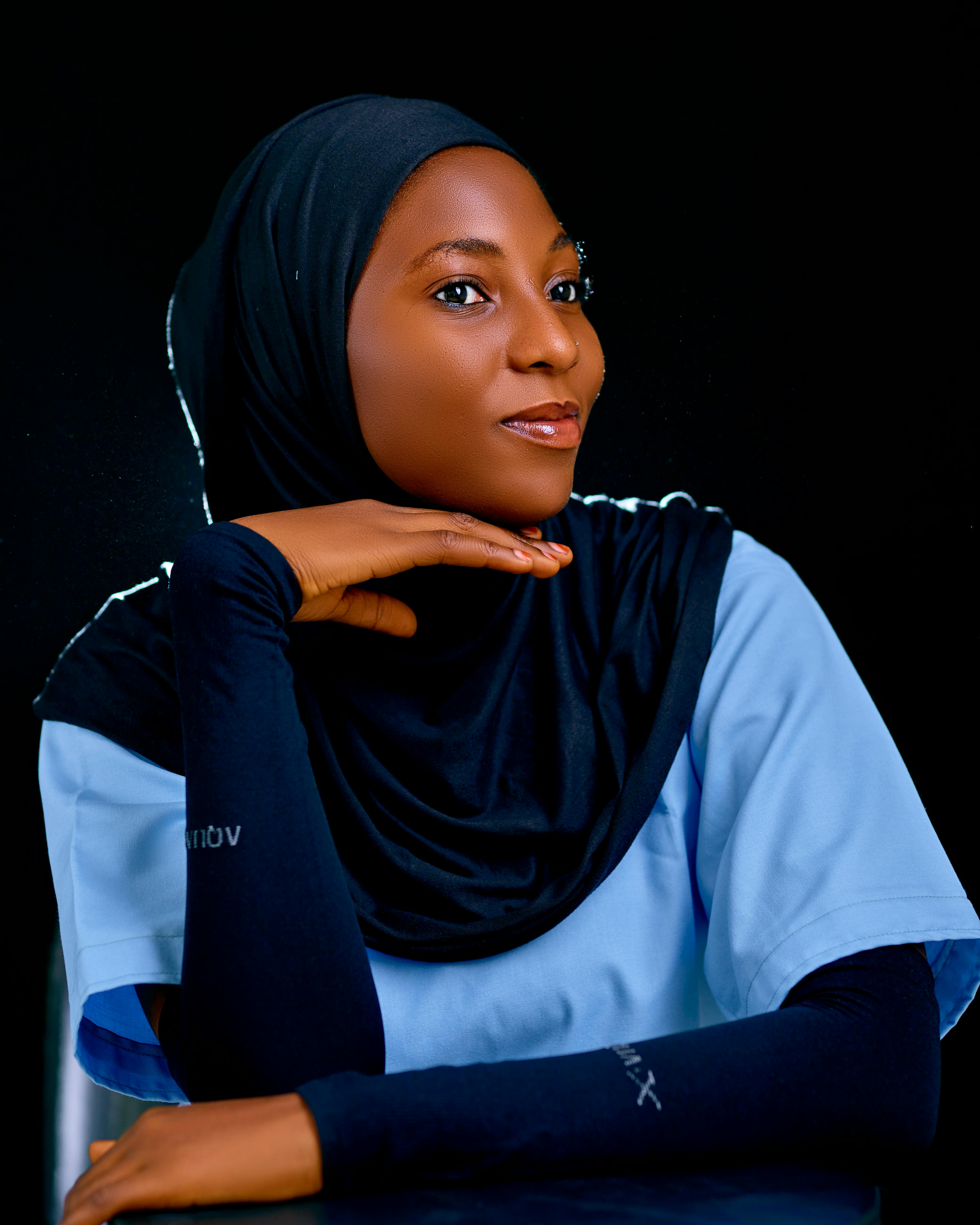 A woman wearing a hijab and medical scrubs, resting her chin on her hand in a thoughtful pose.