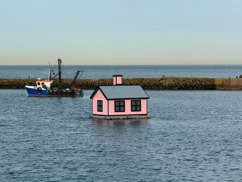 Free A whimsical pink house floating on calm ocean waters with a boat nearby. Stock Photo