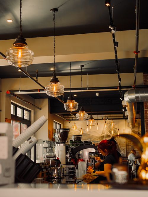 Free Warm cafe interior with industrial lighting and barista at work, creating inviting ambiance. Stock Photo