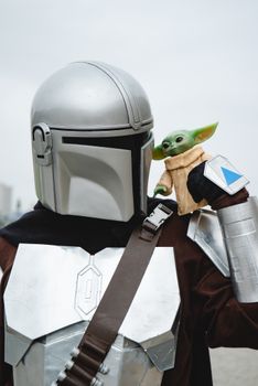Mandalorian cosplayer with Baby Yoda figurine during a sci-fi convention outdoors.