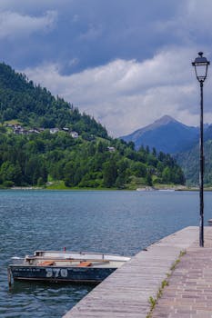 Serene mountain lake with a boat and lantern on the pier, surrounded by lush greenery and hills.