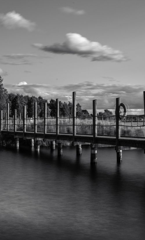 Free A tranquil black and white photo of a lakeside pier capturing serene water and dramatic clouds. Stock Photo