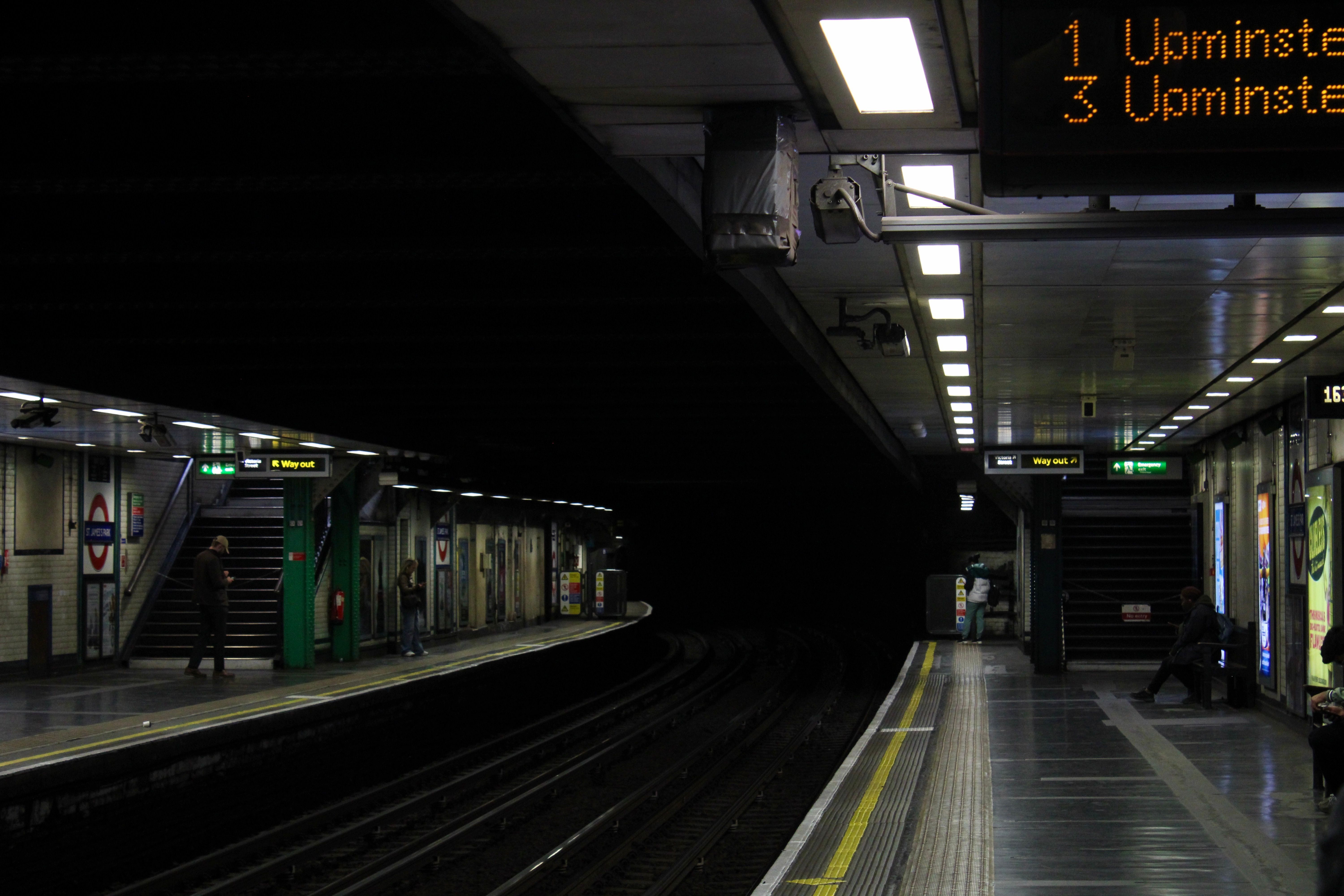 Dimly lit London Underground platform at night with passengers waiting.