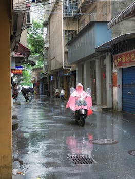 A rainy day in a narrow urban alleyway with a person on a scooter wearing a pink raincoat.