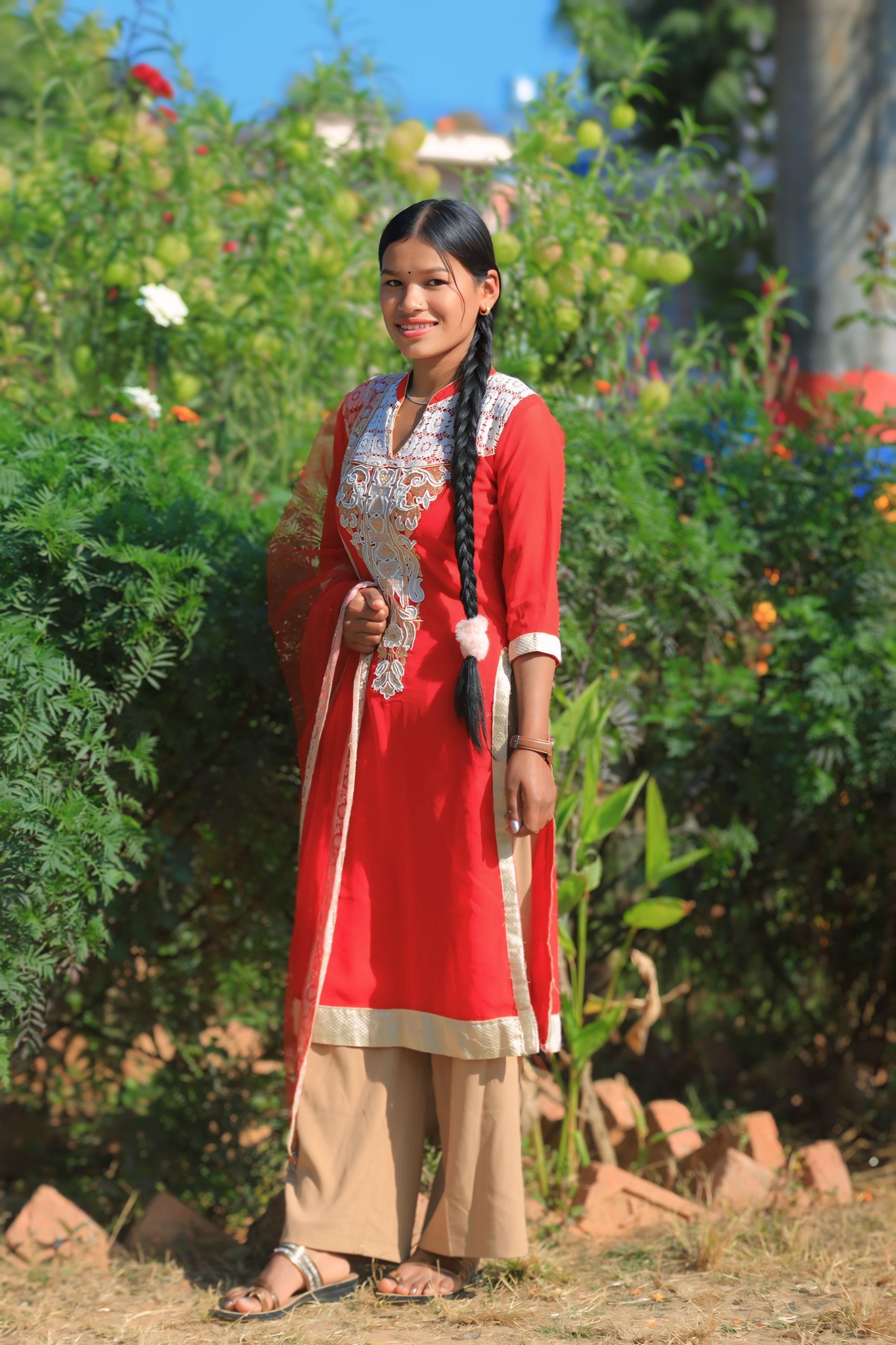 A young woman poses confidently in a red traditional dress surrounded by greenery.