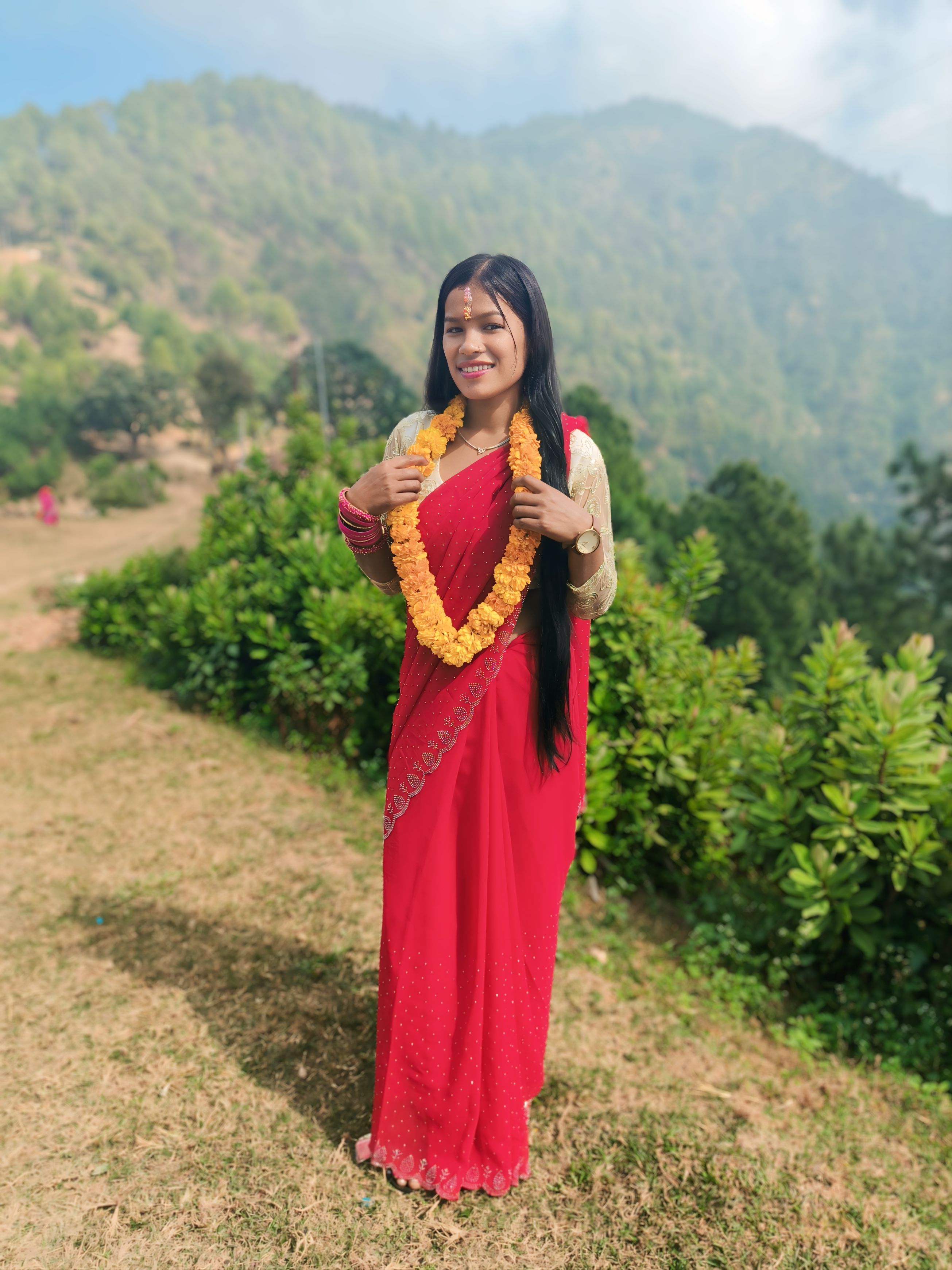 Smiling woman in traditional red saree and marigold garland in lush outdoor setting.