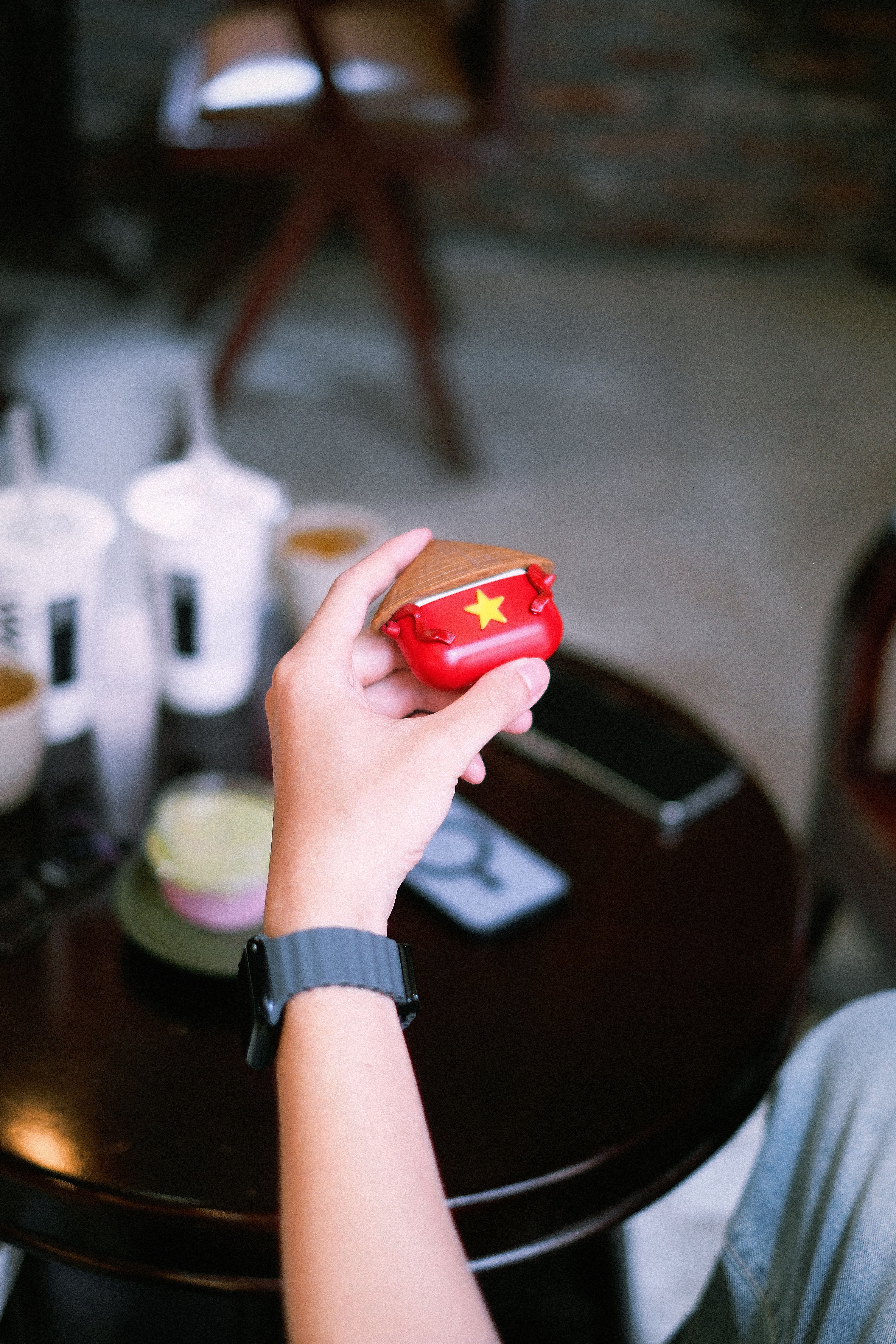 A person holds an AirPods case with a Vietnam flag design in a cafe setting.