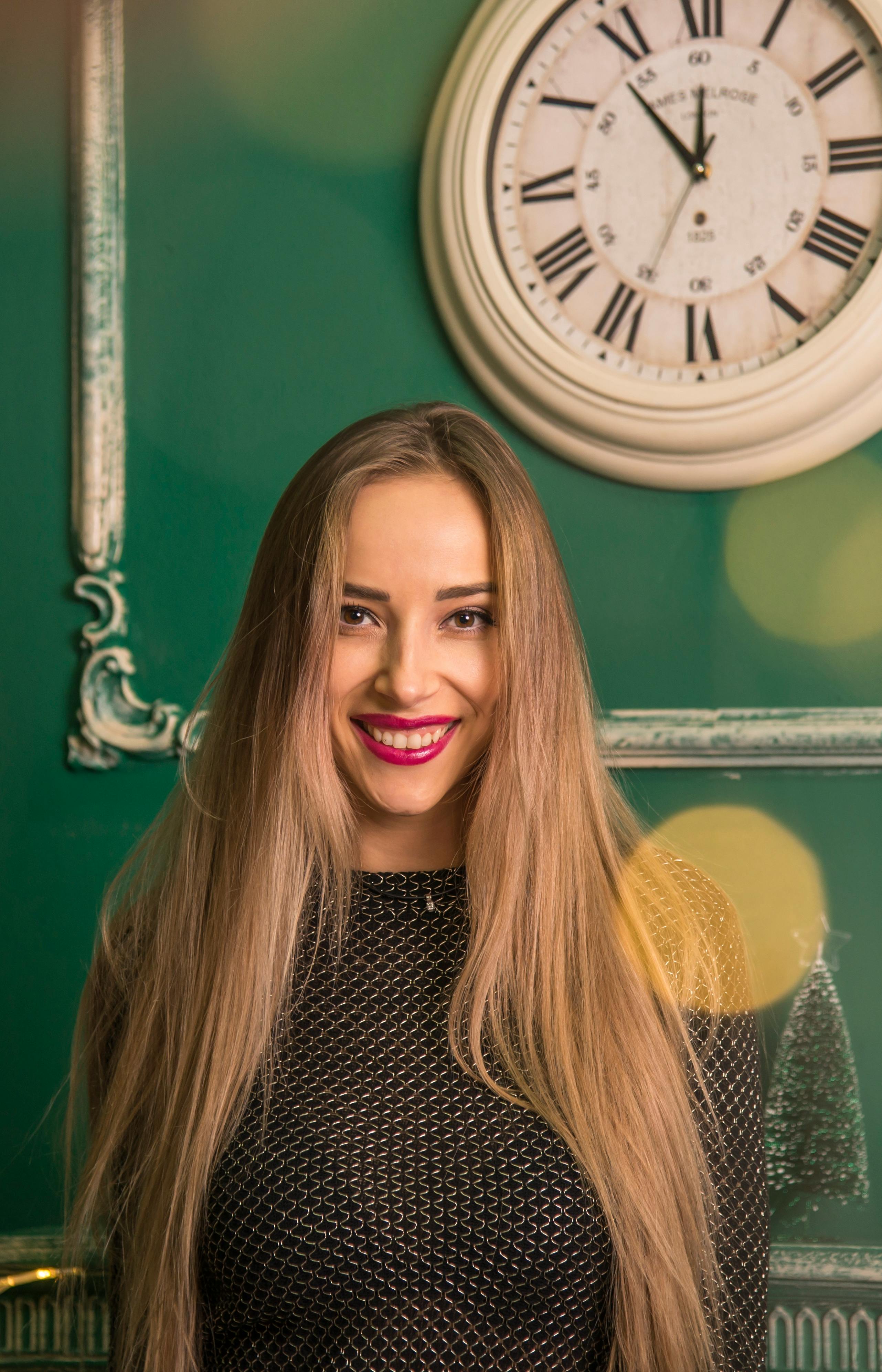 Gratis Una donna dai capelli lunghi sorride radiosamente in casa, con lo sfondo di un orologio d'epoca e di una parete verde. Foto a disposizione