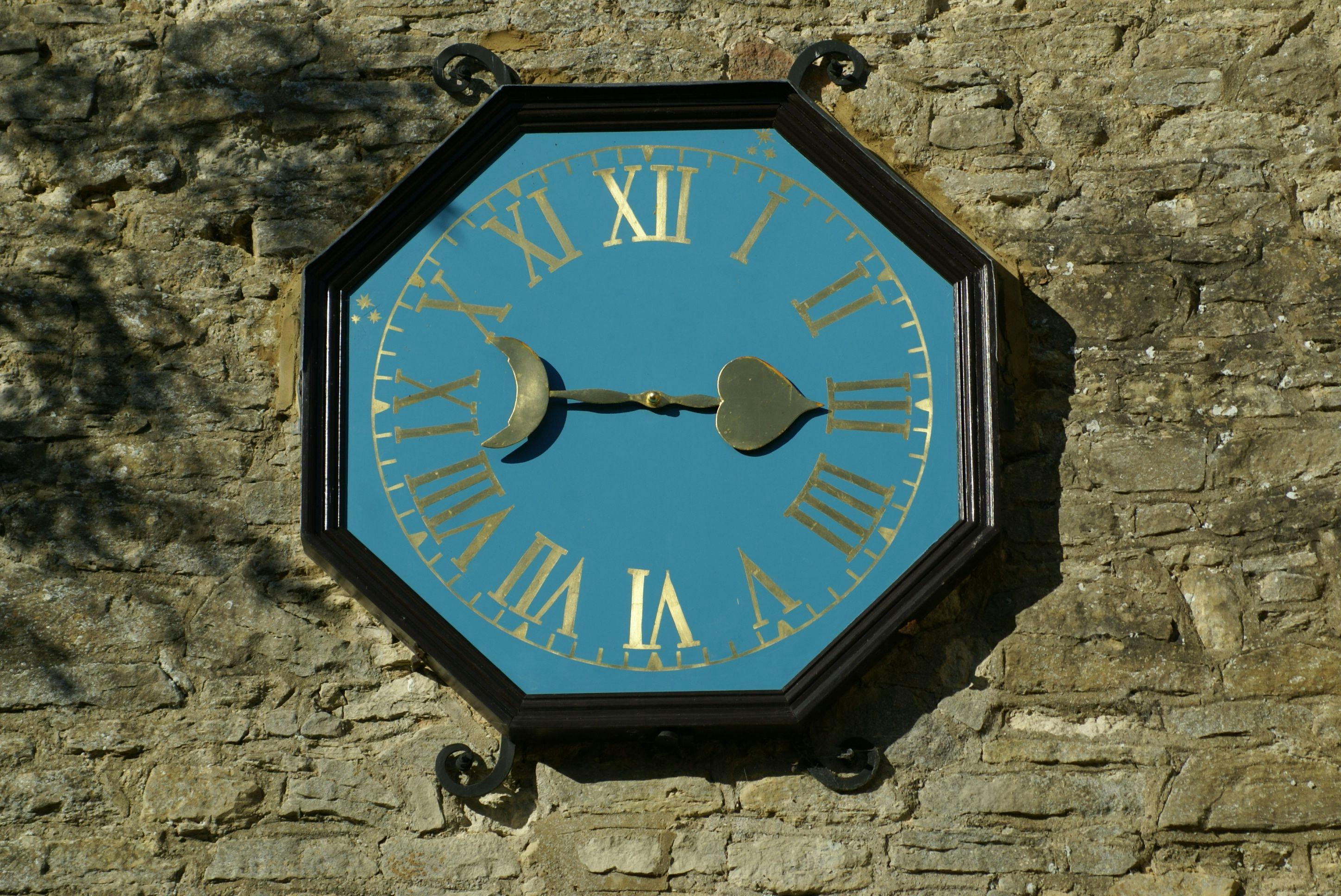 Large vintage clock with Roman numerals mounted on a stone wall in daylight.