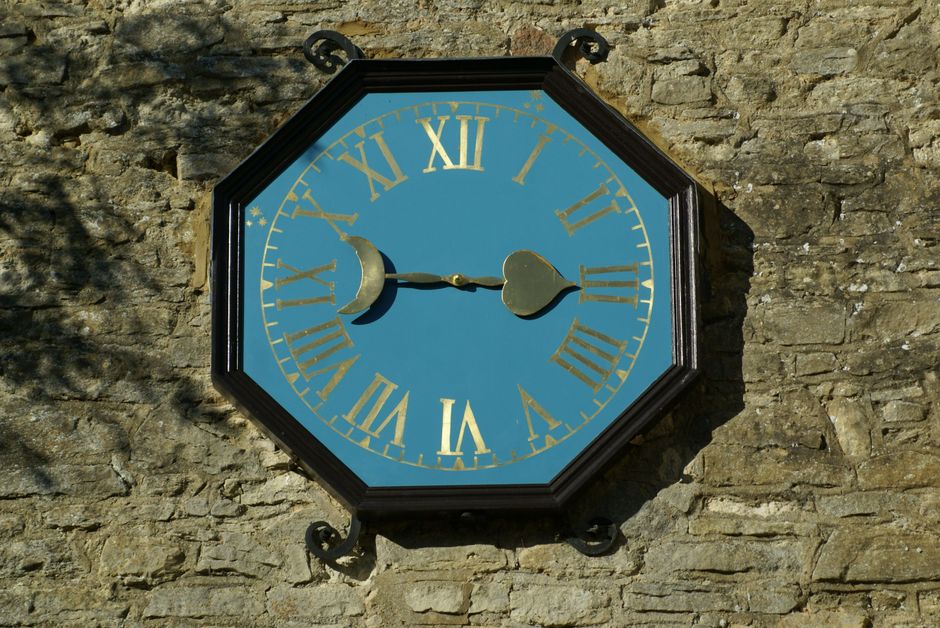 Large vintage clock with Roman numerals mounted on a stone wall in daylight.