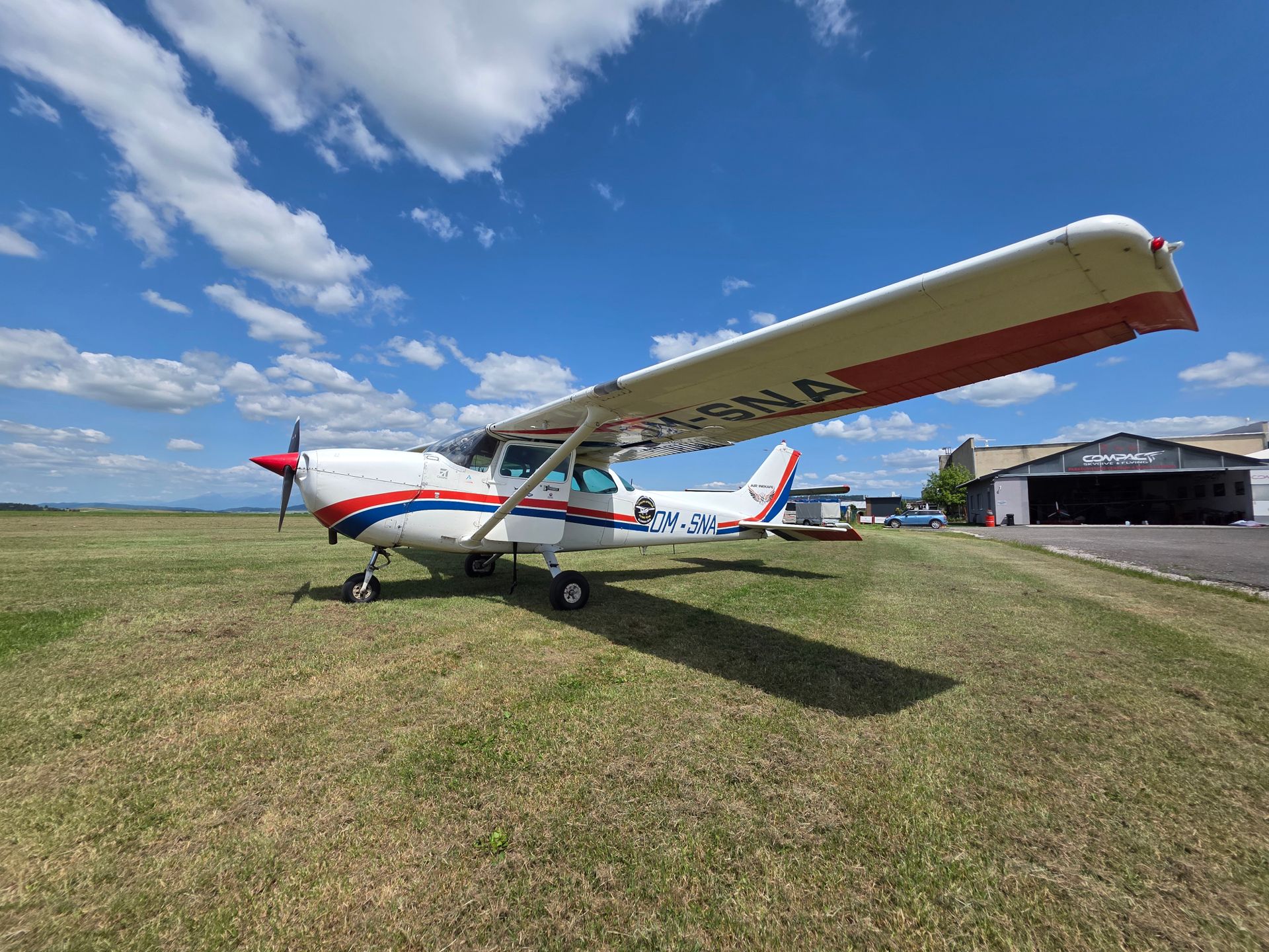 A small aircraft parked on a grassy airfield with a hangar in the background under a clear blue sky.