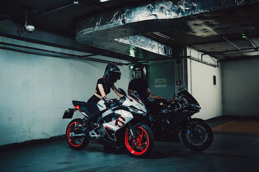 Two motorcycle riders, wearing helmets, are sitting on sports bikes in a dimly lit underground parking garage.
