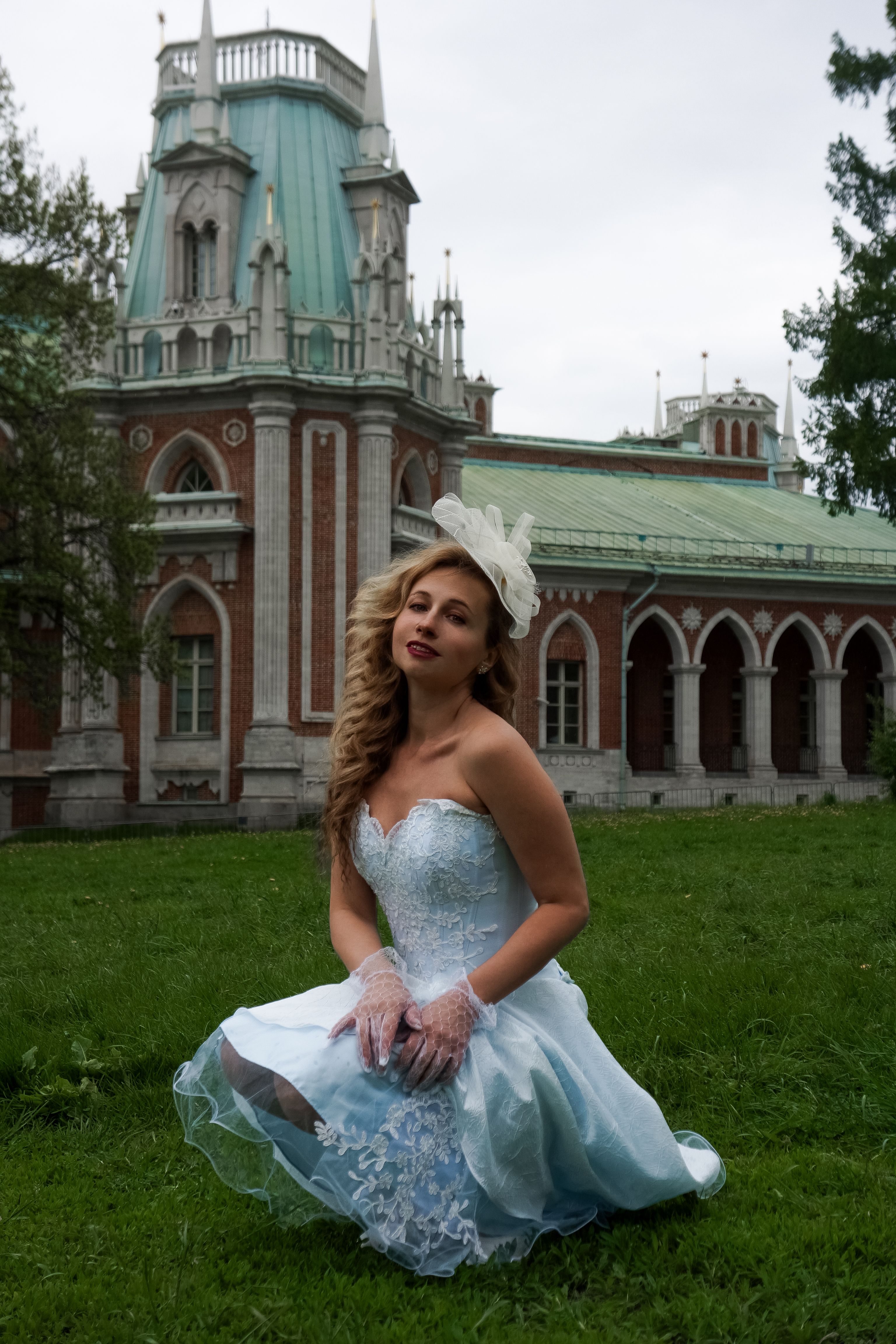 Woman in a blue dress poses elegantly in front of a historic architectural building on a cloudy day.