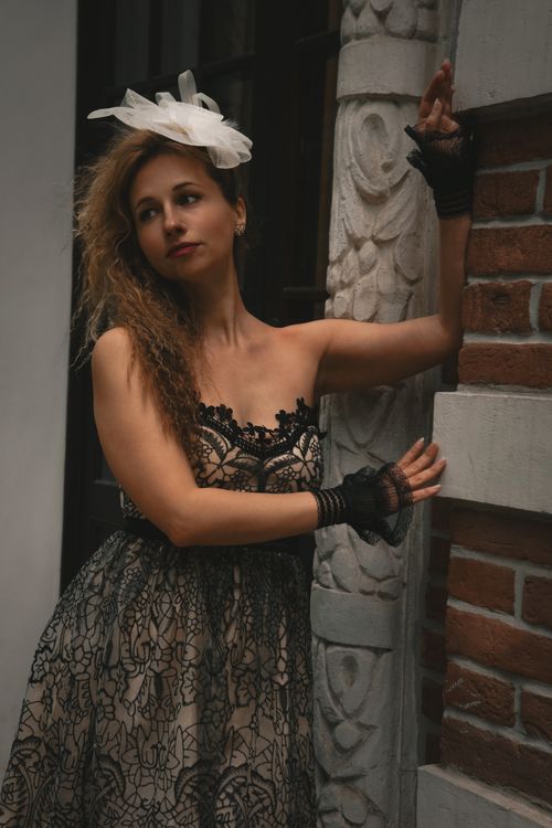 Free Stylish woman in a black lace dress posing near a brick wall with carved details, showcasing elegance and vintage charm. Stock Photo